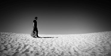 Man walking alone on sand dunes holding a guitar in black and white.
