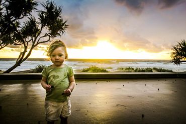 A toddler stands by the beach at sunset with a thoughtful expression.