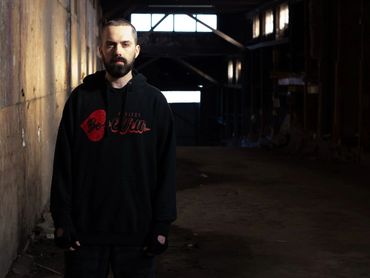 Man with a beard wearing a black hoodie in a dimly lit industrial space.