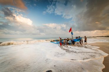 Group of people pushing a blue boat into the ocean on a cloudy day.