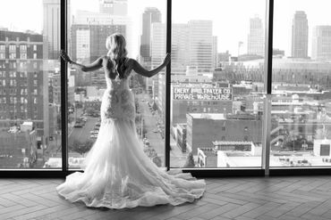 Bride in a flowing gown gazes out over a city skyline through large windows.