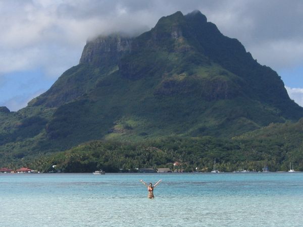 A person wades in clear water near a tropical island with a cruise ship and mountain in the background.