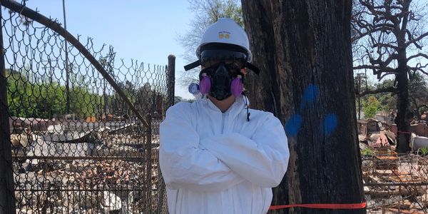 Person in protective suit and respirator at a fire-damaged site.