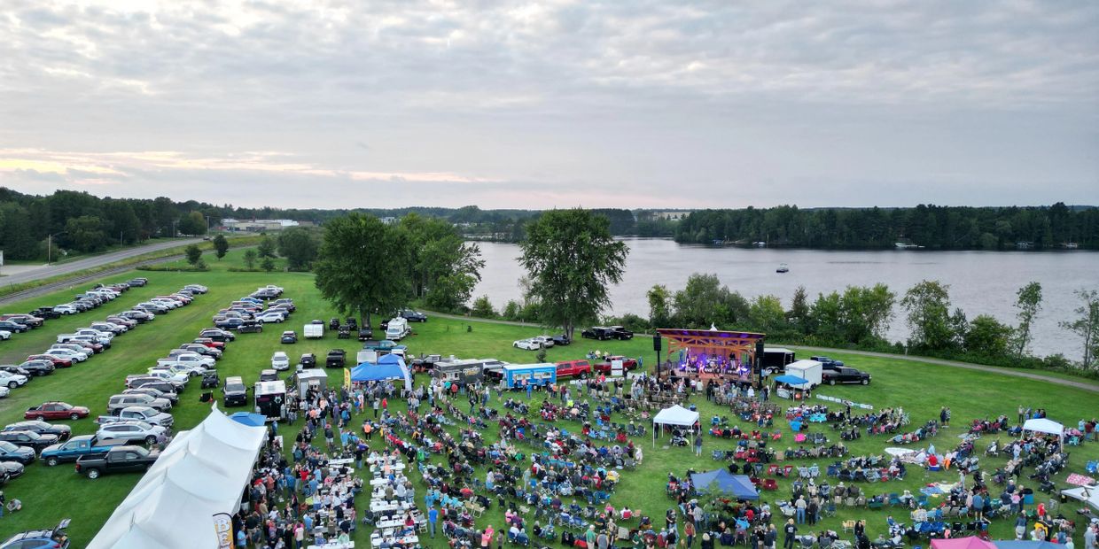Outdoor concert by a lakeside with a large crowd and parked cars on green grass.