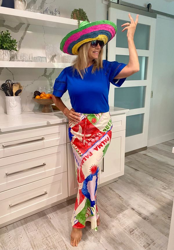 Woman in colorful sombrero, blue top, and vibrant skirt poses in a modern kitchen.