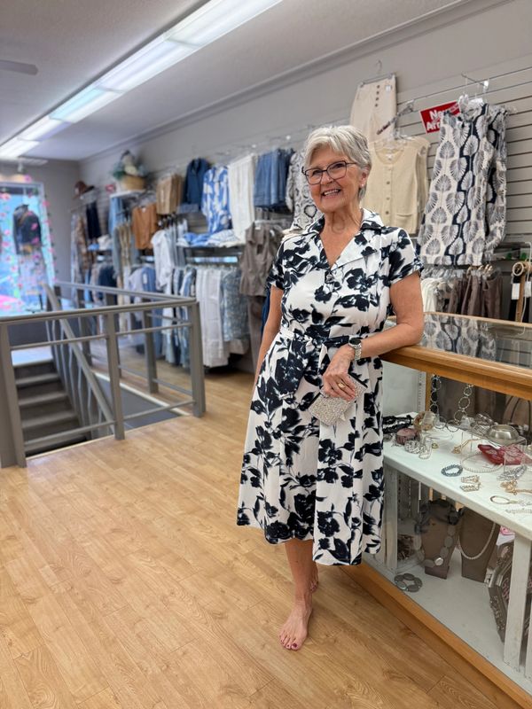 An elderly woman in a black and white floral dress stands barefoot inside a boutique.