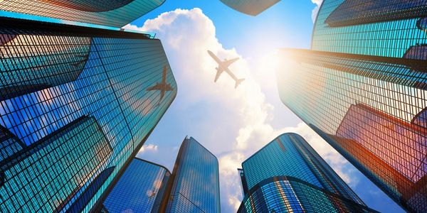 Airplane flying above modern skyscrapers against a bright sky.
