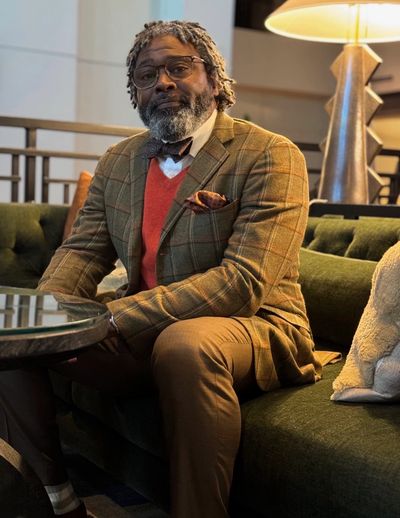 Man in a stylish brown checkered blazer and red sweater sitting on a green couch.