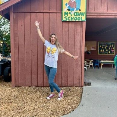 Woman excitedly posing outside a small wooden school building.