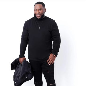 Smiling man in black outfit holding a jacket against a white background.