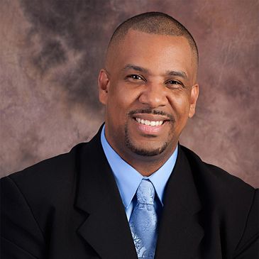 Smiling man in a black suit and blue tie against a brown backdrop.
