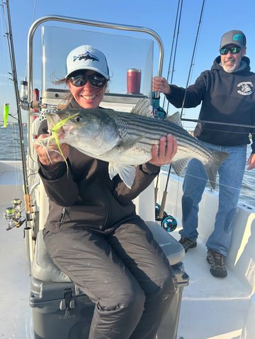 Woman proudly holding a large striped bass on a boat with a smiling man standing behind.