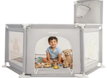 Toddler playing inside a gray playpen with toys and a teddy bear.
