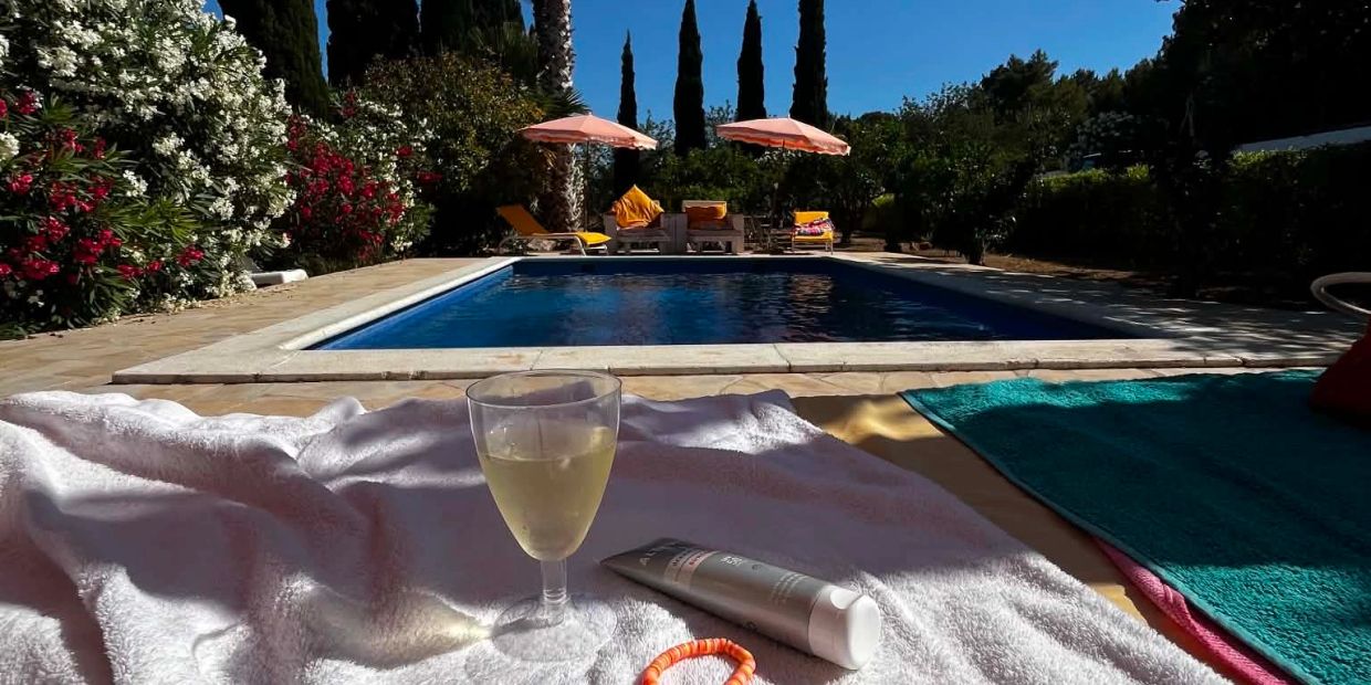 Relaxing poolside scene with a drink, sunscreen, and umbrellas under a clear blue sky.