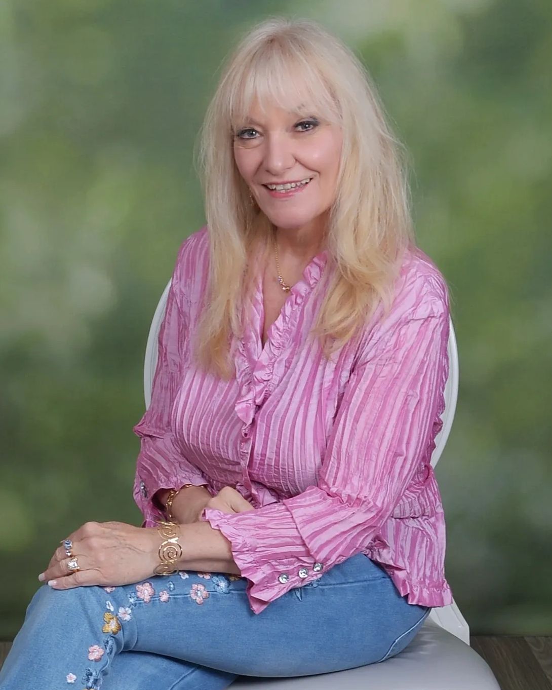 Smiling woman in a pink blouse and floral jeans seated on a white chair.