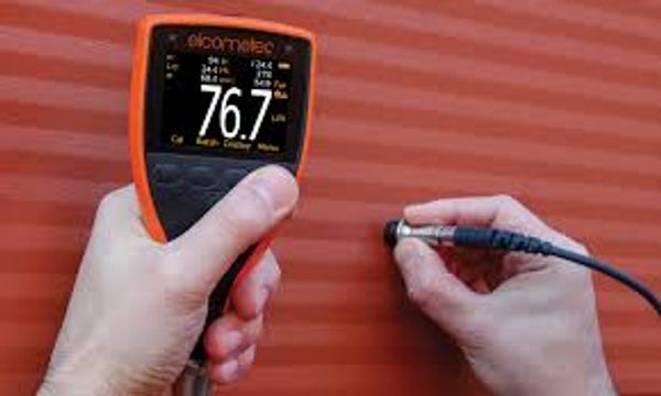 Handheld coating thickness gauge measuring on a red surface.
