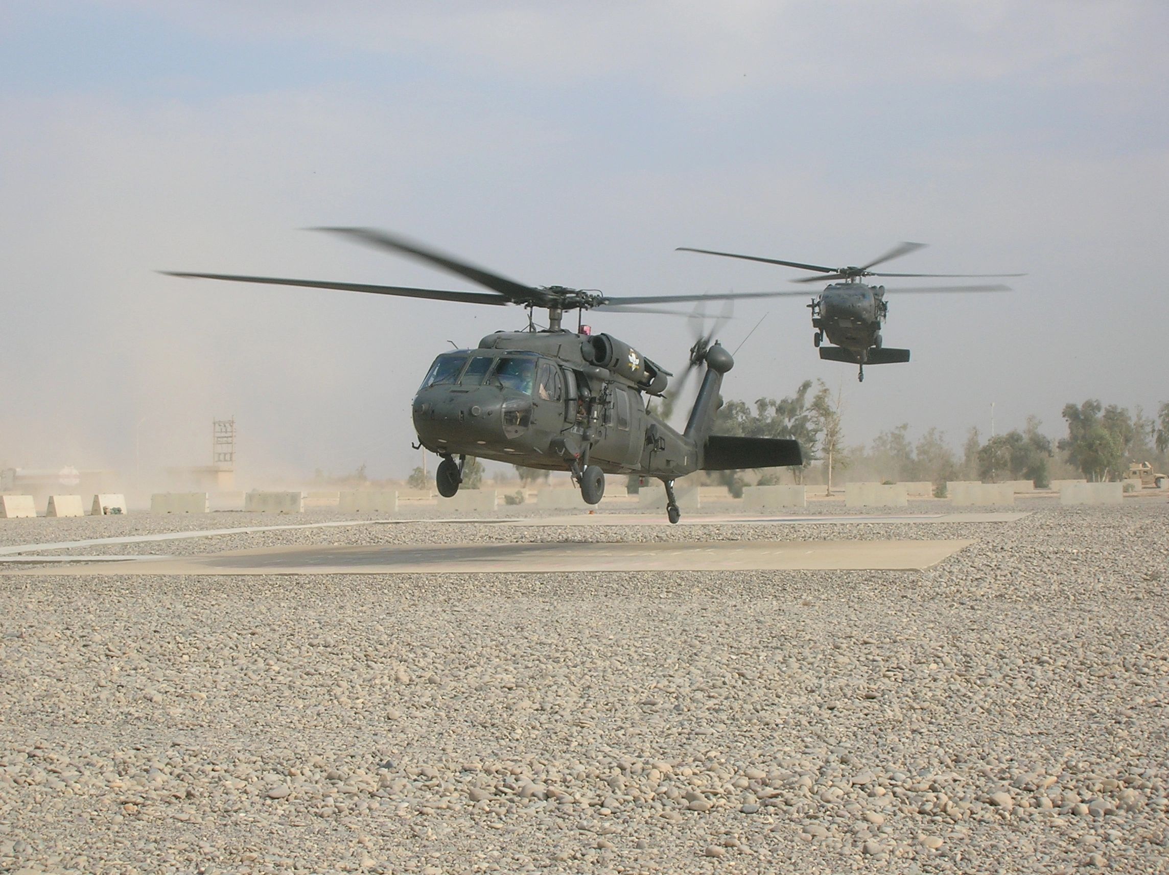 Two military helicopters in flight near a desert airstrip.