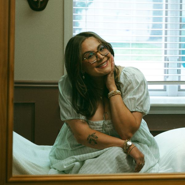 A smiling woman in glasses and a light dress sits comfortably indoors.