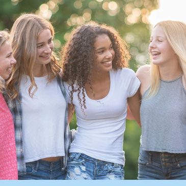A group of five teenage girls smiling and embracing outdoors in casual summer clothes.