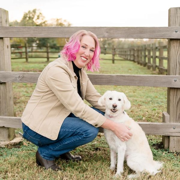 Woman with pink hair crouches beside a happy white dog by a wooden fence.