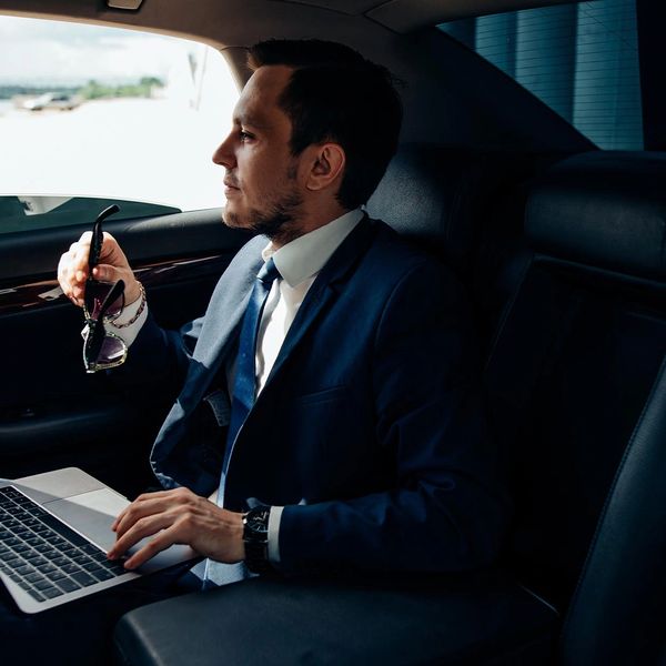 Businessman working on laptop inside a luxury car, holding sunglasses.