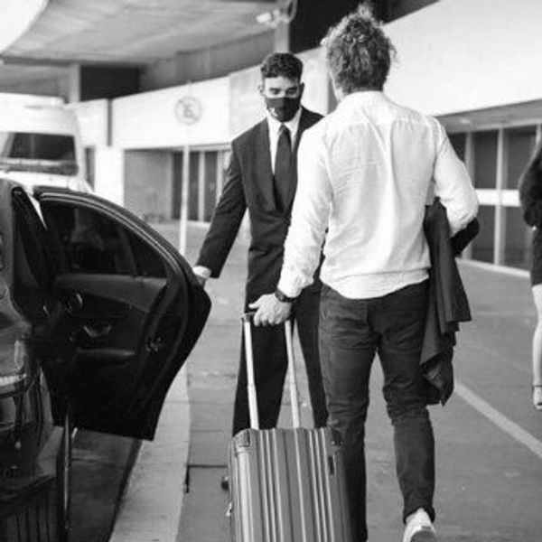 A man with luggage is greeted by a chauffeur near a luxury car at an airport.