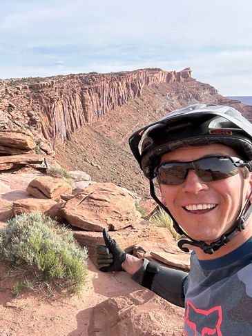 Smiling cyclist giving thumbs up on rocky desert trail with cliffs behind.
