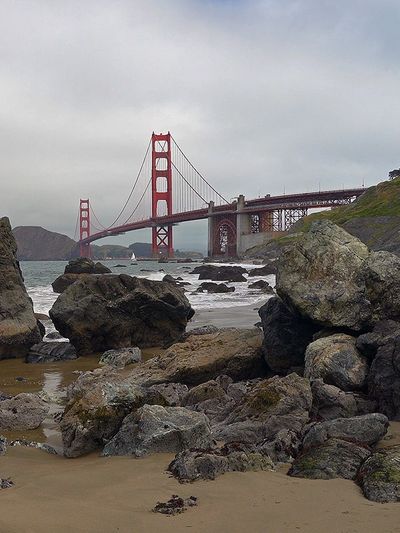 Golden Gate Bridge viewed from rocky beach under cloudy sky.