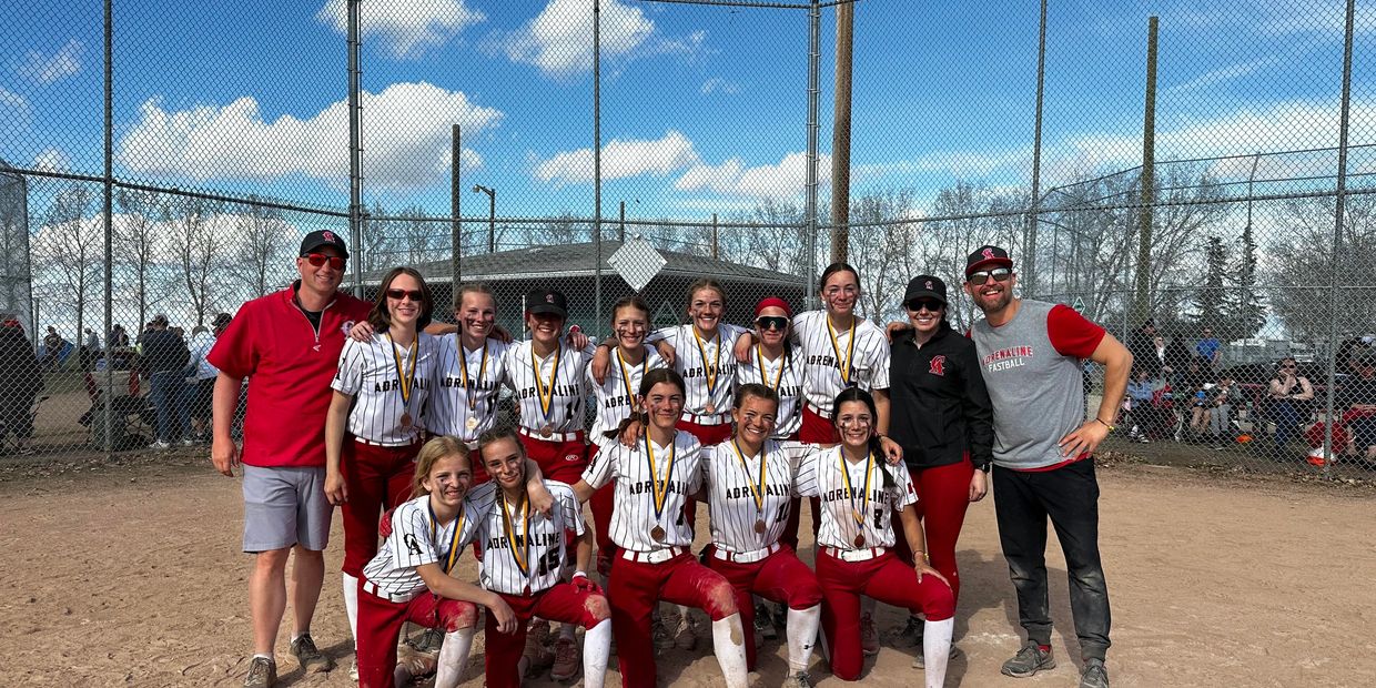Youth softball team posing with medals on a sunny day at the field.