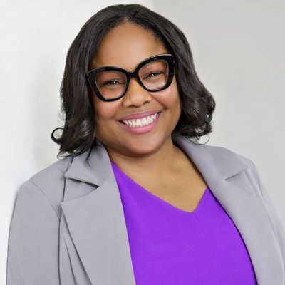 Smiling woman wearing large black glasses and a purple top with a gray blazer.