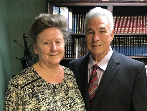 An elderly couple posing together indoors with bookshelves in the background.
