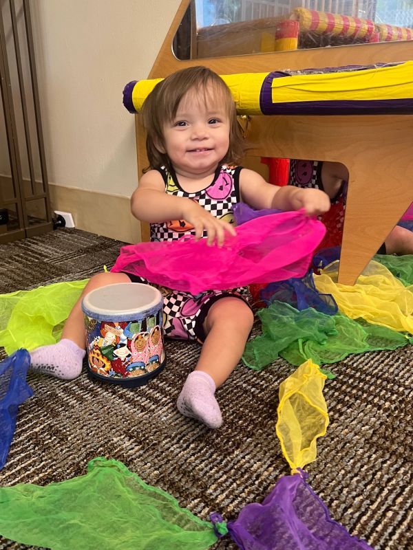 Smiling toddler playing with colorful scarves and drum.