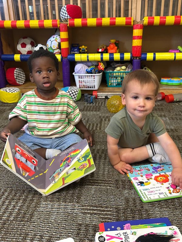 Two toddlers reading colorful books while sitting on the floor.