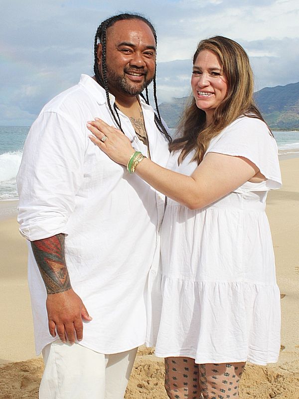 Smiling couple in white outfits posing on a beach with ocean and mountains in the background.