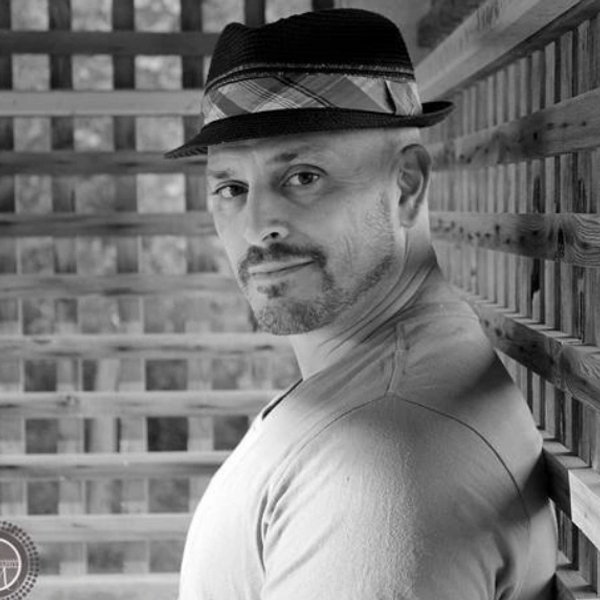 Man in a hat leans against a wooden lattice wall, looking at the camera.