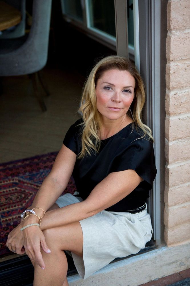 Woman with blonde hair sitting by a doorway, wearing a black top and light skirt.