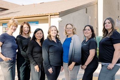Seven women standing outdoors in casual attire, smiling for a group photo.
