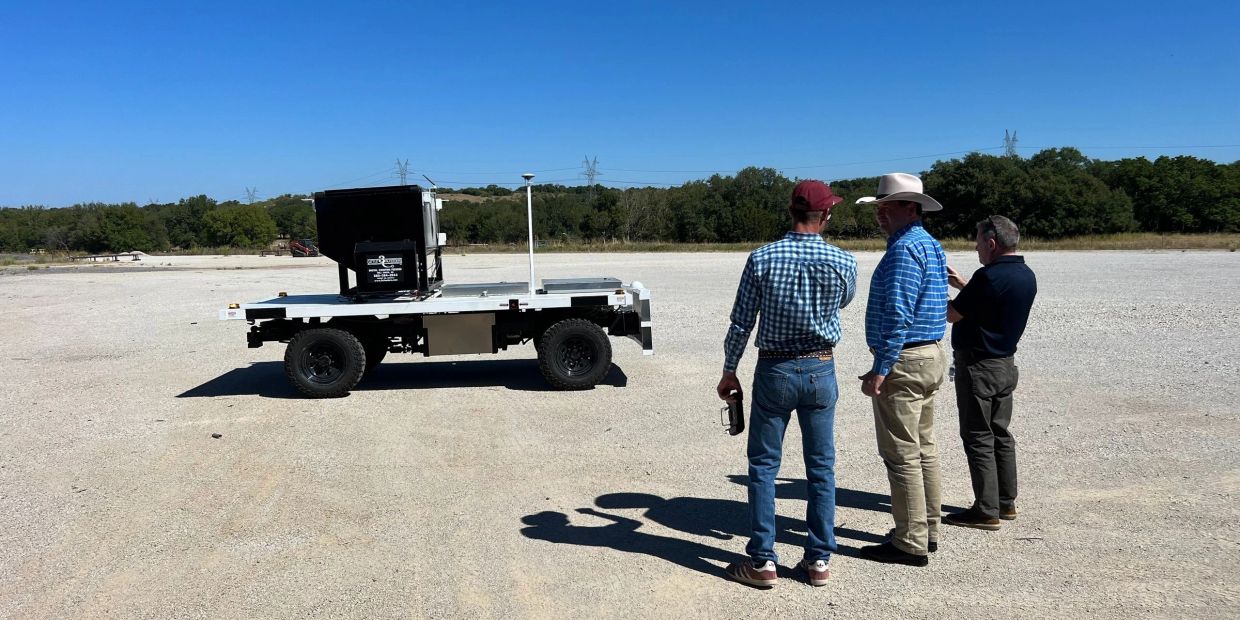 Three men observe a robotic vehicle on a gravel lot under a clear blue sky.