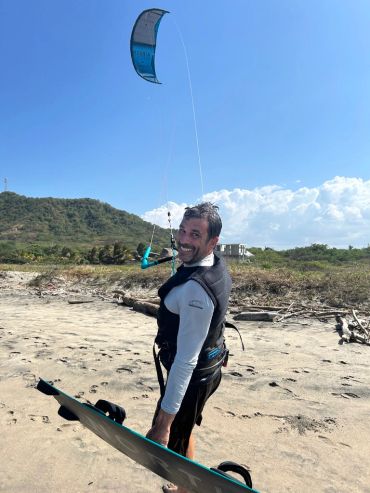 Un homme souriant avec un équipement de kitesurf sur la plage.