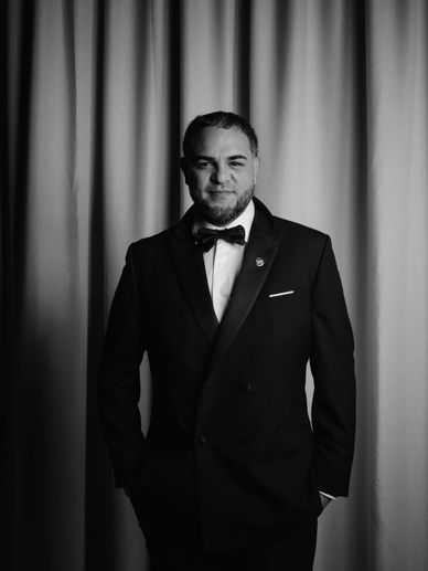 Man in a tuxedo standing in front of curtains, smiling.
