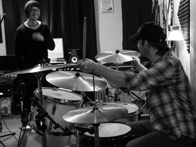 Two musicians in a recording studio, one playing drums and the other smiling.