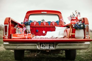 Red pickup truck bed decorated for a patriotic celebration with flags and cushions.