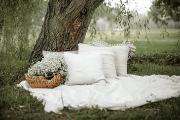 Cozy outdoor picnic setup with white pillows and a basket of flowers under a tree.