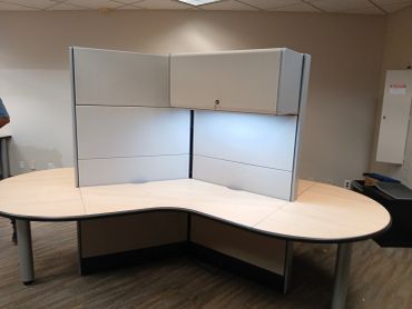 Empty office cubicle with wooden desk and overhead cabinets.