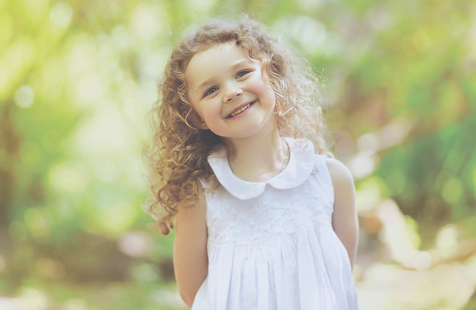 Une jeune fille avec une robe blanche et les cheveux frisés sourit dans la forêt.