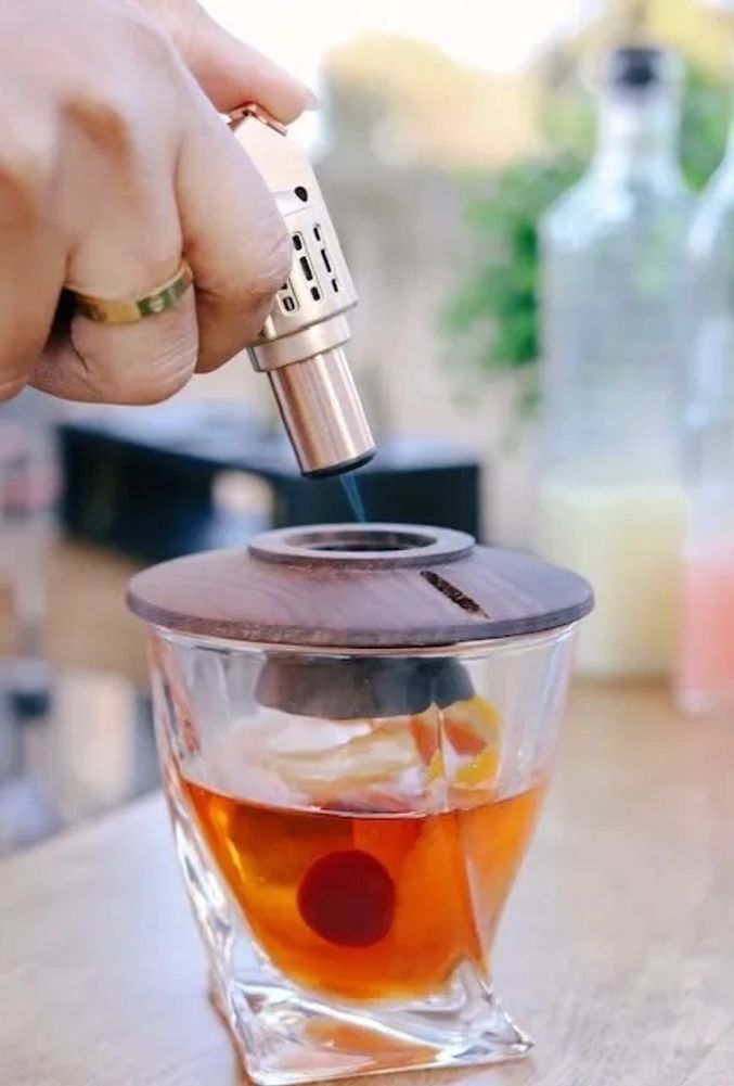 A hand uses a torch lighter to ignite a wooden cover on a glass with a drink.
