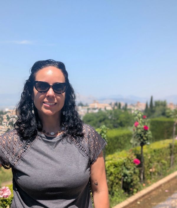 Woman with sunglasses smiling outdoors by a garden.