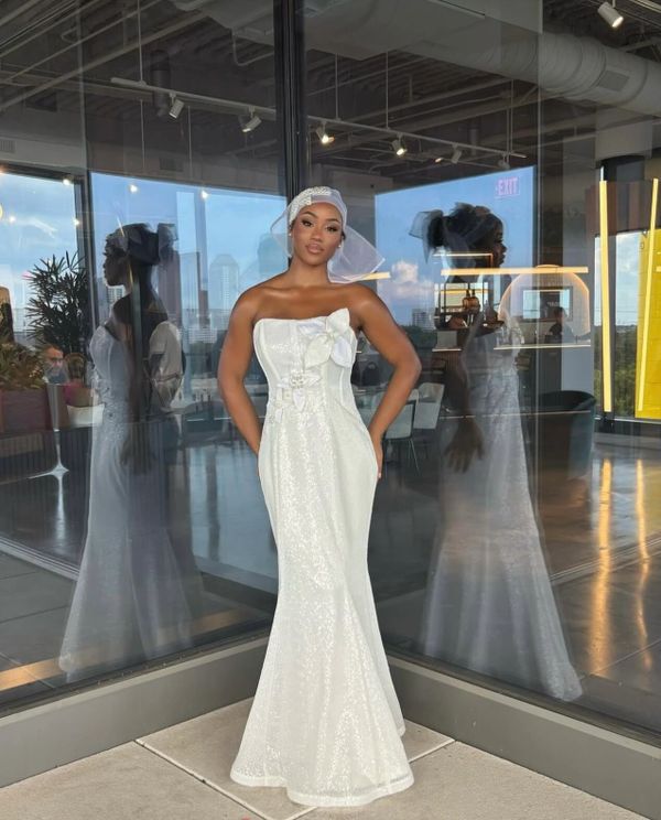 Woman in elegant white strapless gown posing by glass walls.