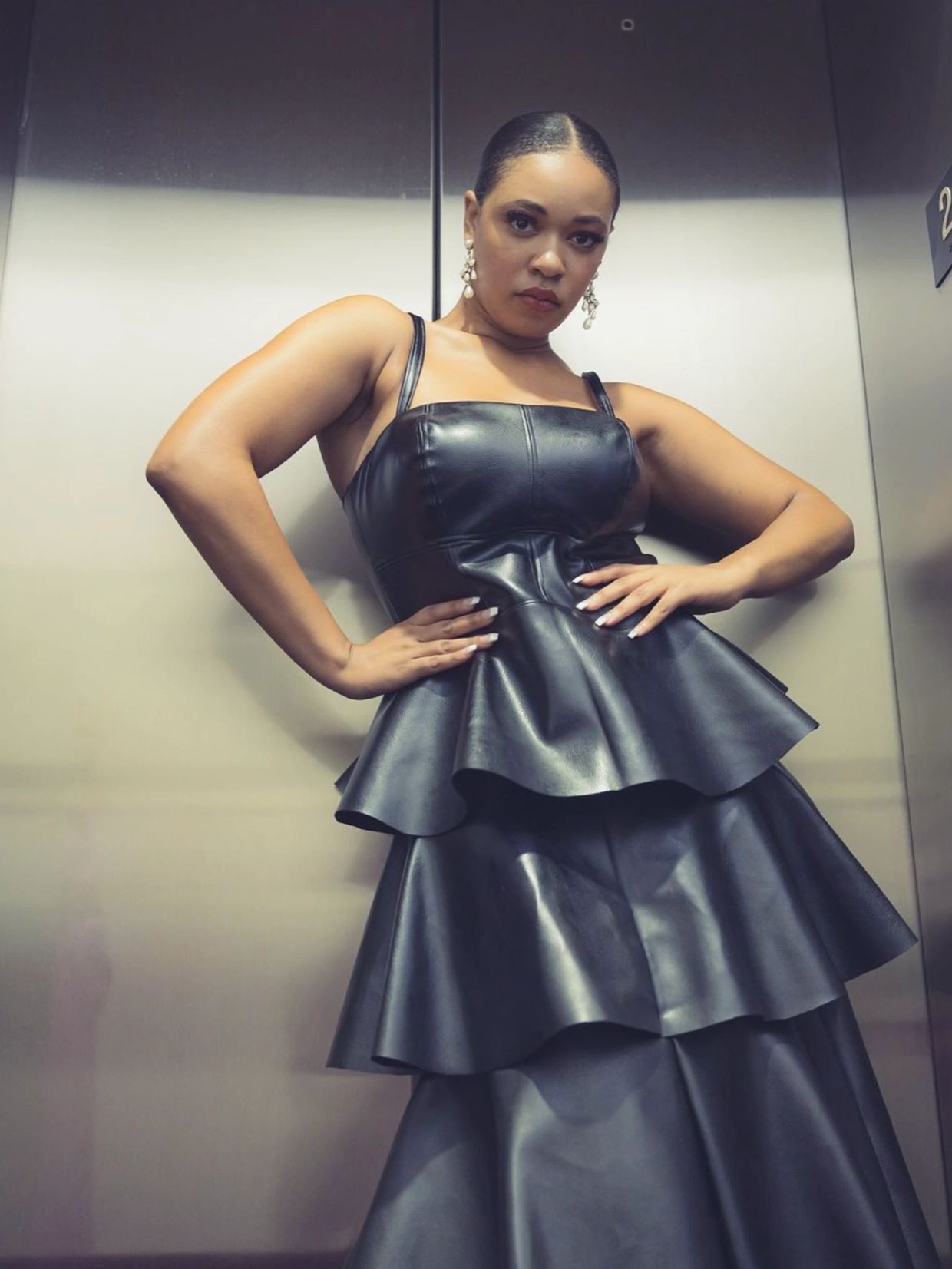 Woman posing confidently in a black leather tiered dress with pearl earrings.
