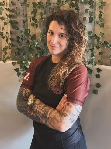 Woman with curly hair and tattoos, arms crossed, smiling gently.